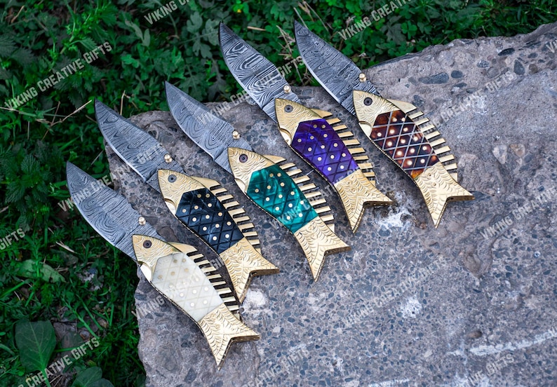 May include: Six folding pocket knives with fish-shaped handles. The knives have Damascus steel blades and handles made of different colored materials, including purple, green, black, and white.