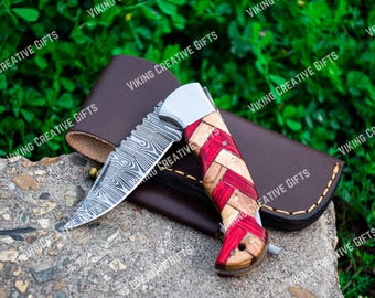 Puede incluir: Un cuchillo plegable de bolsillo con una hoja de acero Damasco y un mango de madera rojo y blanco. El cuchillo está apoyado en una funda de cuero marrón.
