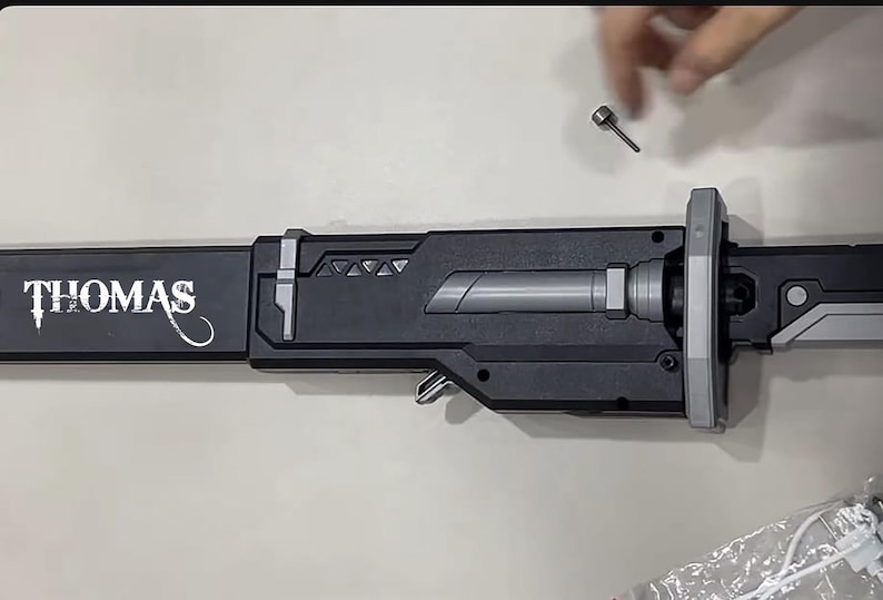 May include: A black and gray sword with the name "THOMAS" in white lettering. The sword has a silver handle and a silver blade. A small silver screw is above the sword.