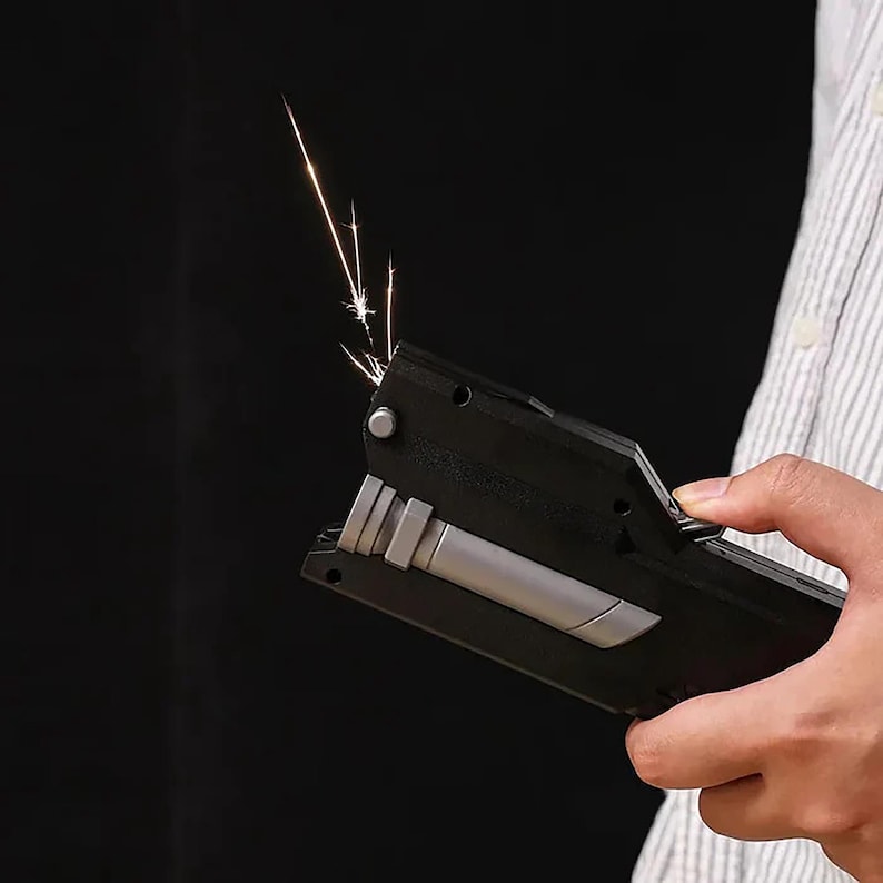 May include: A black and silver lighter with sparks emitting from the top. The lighter is held in a person's hand against a dark background. The lighter has a rectangular shape with a silver accent.