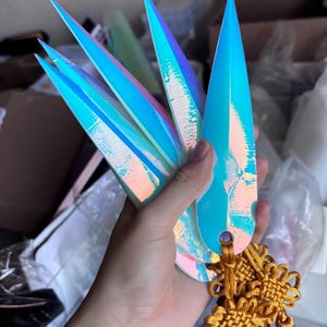May include: A set of iridescent, pointed nail tips in shades of blue, purple, and green. The tips are held together with a gold-colored decorative element. The nails have a sharp, elongated shape.