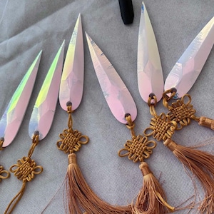 May include: Six iridescent, teardrop-shaped pendants with gold-colored knot accents and long, brown tassels. The pendants have a pink and white color scheme and are arranged on a gray fabric surface.