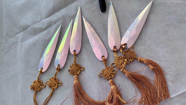 May include: Six iridescent, teardrop-shaped pendants with gold-colored knot accents and long, brown tassels. The pendants have a pink and white color scheme and are arranged on a gray fabric surface.