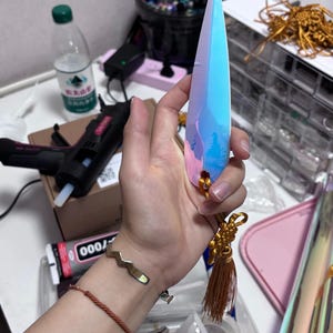 May include: A long, iridescent crystal-shaped object with blue, pink, and purple hues. It has a gold tassel and a small gold accent at the base. The object is held in a hand, with a gold bracelet visible on the wrist.