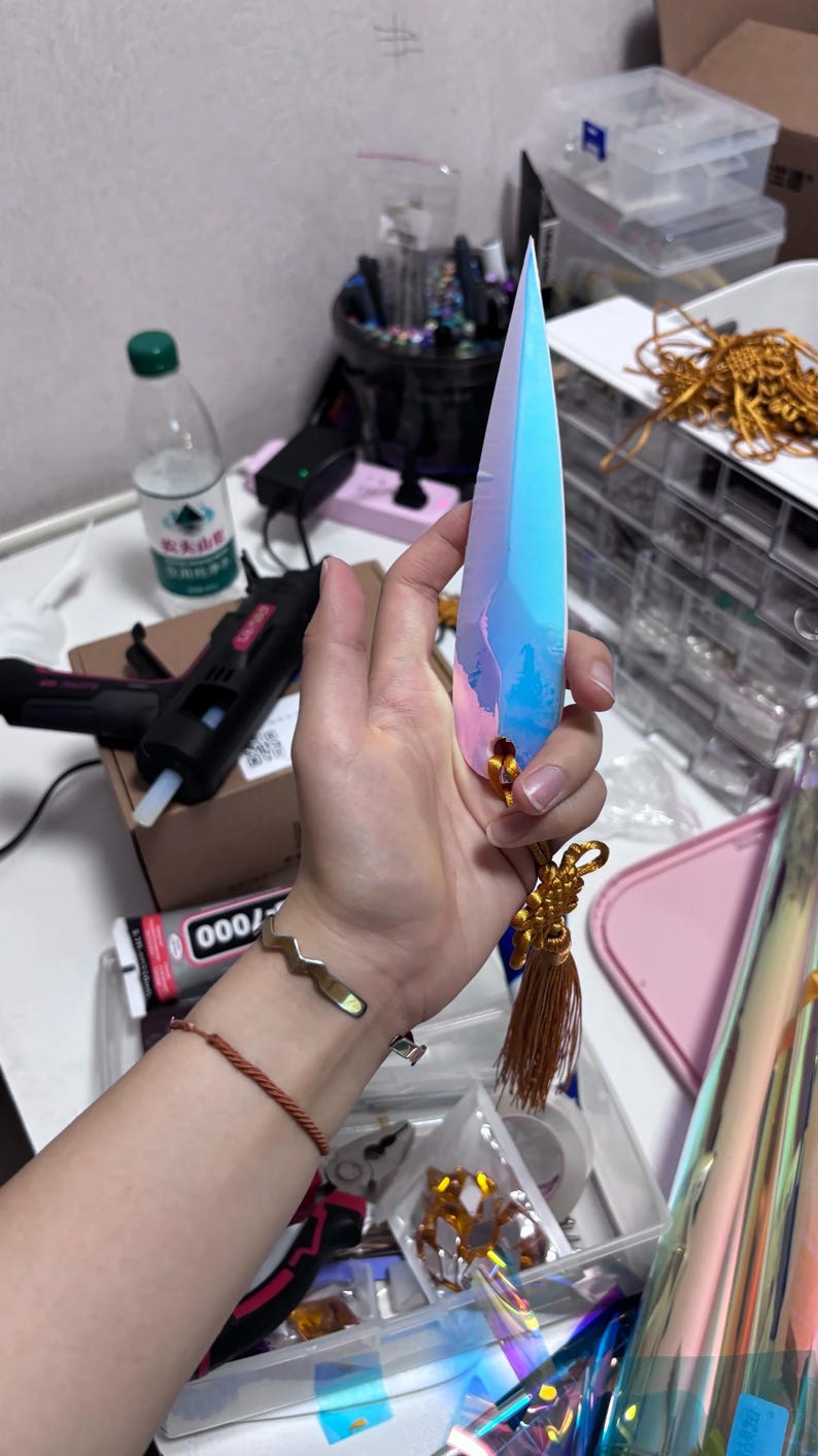 May include: A long, iridescent crystal-shaped object with blue, pink, and purple hues. It has a gold tassel and a small gold accent at the base. The object is held in a hand, with a gold bracelet visible on the wrist.