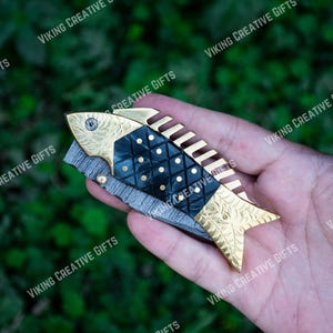 May include: A gold and black fish-shaped pocket knife with a decorative pattern. The knife has a serrated blade and a comb on the back.