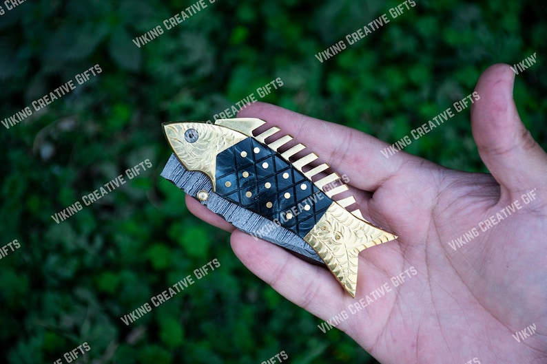 May include: A gold and black fish-shaped pocket knife with a decorative pattern. The knife has a serrated blade and a comb on the back.