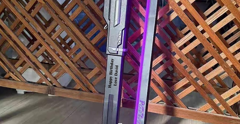 May include: A gray and silver sword with a purple light strip. The sword has the text "Happy Birthday Love David" engraved on the side. The background is a wooden lattice.