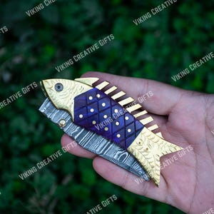 May include: A gold and purple fish-shaped pocket knife with a Damascus steel blade. The knife is open and the blade is visible.
