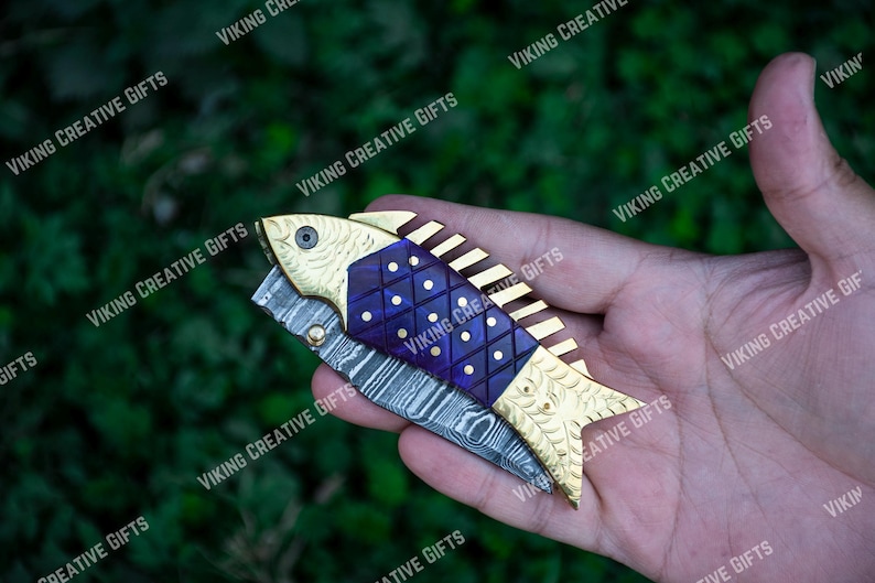 May include: A gold and purple fish-shaped pocket knife with a Damascus steel blade. The knife is open and the blade is visible.