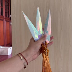 May include: Three iridescent, dagger-shaped objects with a gold tassel. The objects have a pearlescent finish that reflects multiple colors. The objects are held in a hand, with a brown bracelet visible on the wrist.