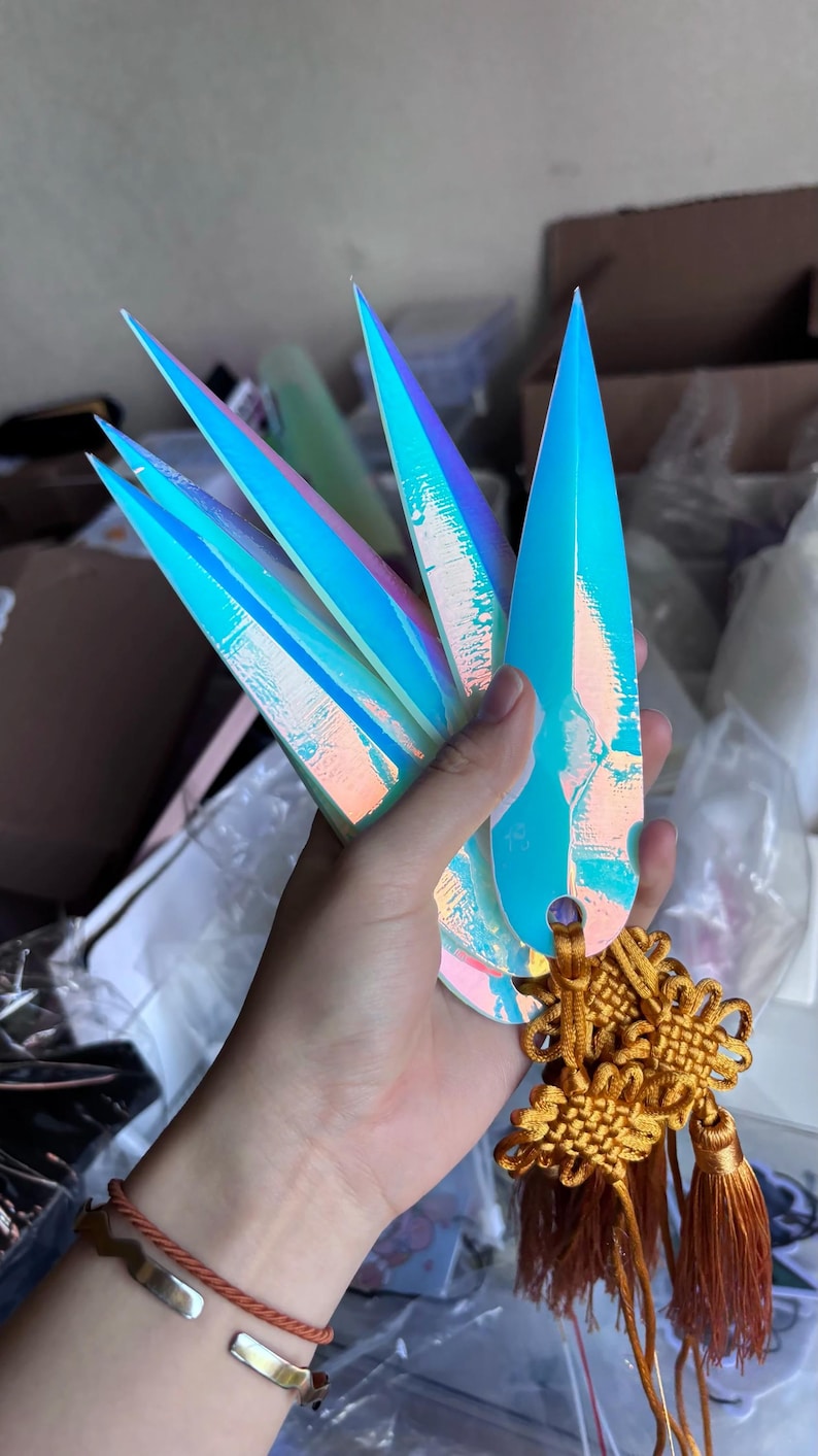 May include: Iridescent blue nail art pieces with sharp, pointed tips. The nail art is held with a gold-colored decorative knot and tassels. The nails have a holographic effect, reflecting multiple colors.