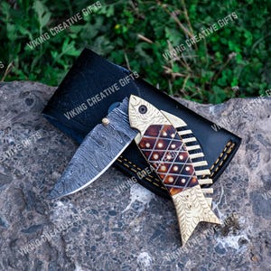 May include: A folding pocket knife with a decorative fish design on the handle. The handle is made of wood and has a gold finish. The blade is made of stainless steel and has a Damascus pattern.