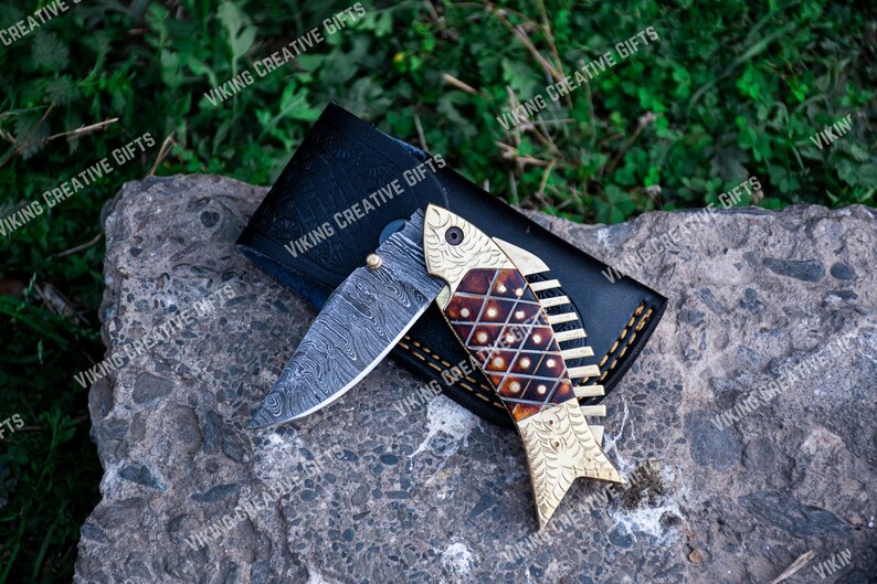 May include: A folding pocket knife with a decorative fish design on the handle. The handle is made of wood and has a gold finish. The blade is made of stainless steel and has a Damascus pattern.