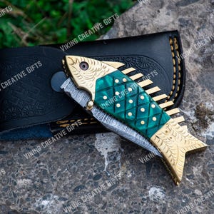 May include: A green and gold pocket knife with a fish design on the handle. The knife is open and the blade is visible. The knife is in a black leather sheath.