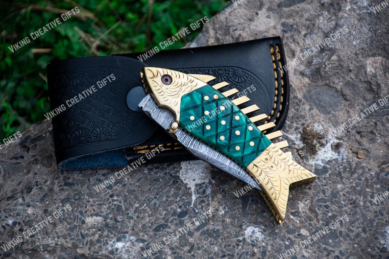 May include: A green and gold pocket knife with a fish design on the handle. The knife is open and the blade is visible. The knife is in a black leather sheath.