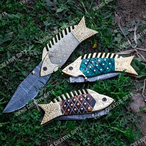 May include: Three decorative folding knives with fish-shaped handles. The handles are made of wood and metal with intricate patterns. The blades are made of stainless steel and have a Damascus pattern.