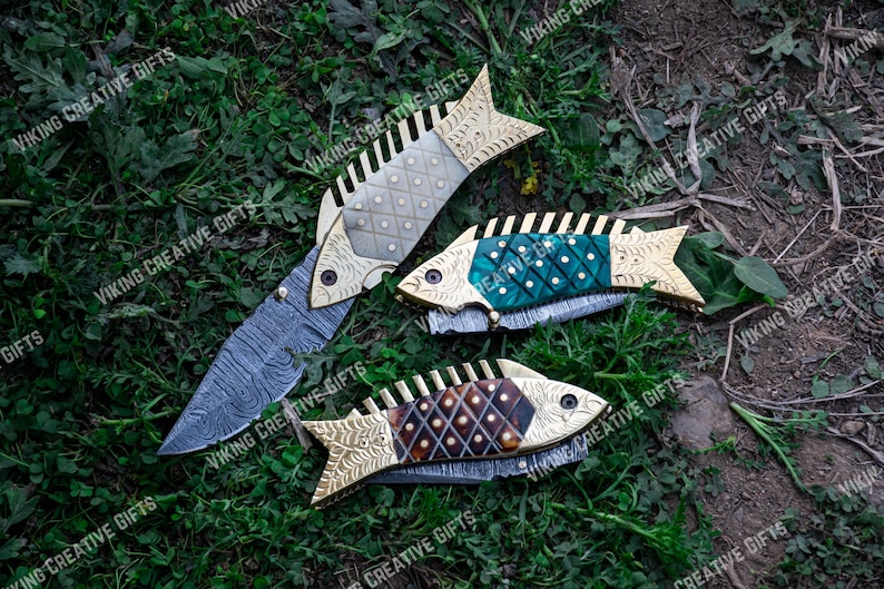 May include: Three decorative folding knives with fish-shaped handles. The handles are made of wood and metal with intricate patterns. The blades are made of stainless steel and have a Damascus pattern.