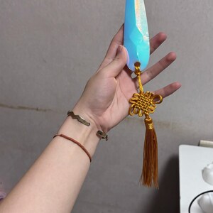 May include: A blue crystal-shaped pendant with a gold tassel and knot detail. The pendant is held by a hand, and the crystal has a faceted design. The tassel is a warm brown color.