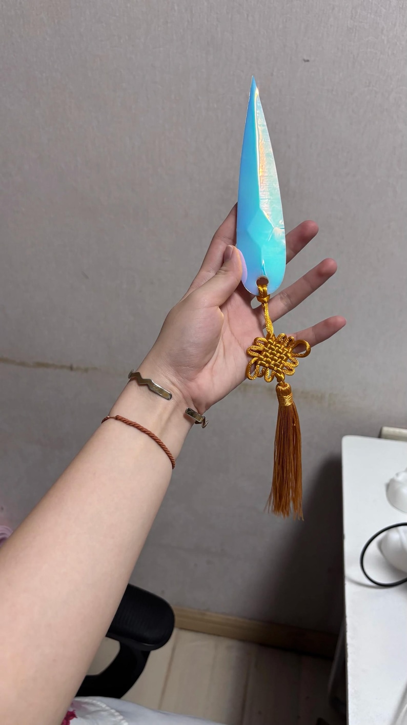 May include: A blue crystal-shaped pendant with a gold tassel and knot detail. The pendant is held by a hand, and the crystal has a faceted design. The tassel is a warm brown color.