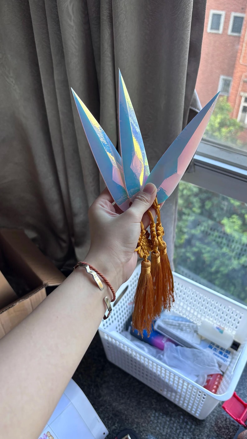 May include: Three iridescent kunai knives with gold tassels are held in a hand. The knives have a sharp, pointed design and reflect a range of colors. The tassels are a warm, golden-orange color.