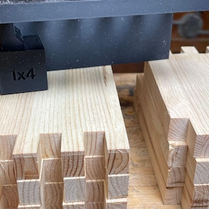 May include: Two stacks of wooden pieces with intricate interlocking joints. The pieces are arranged in a staggered pattern, creating a visually appealing design. A black jig with the label "1x4" is visible above the stacks.