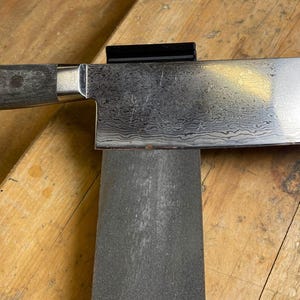 Op de afbeelding: Een close-up van een zilveren keukenmes met een houten handvat dat wordt geslepen op een grijze slijpstaal.