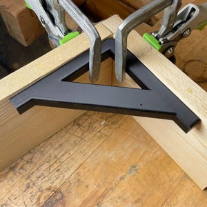 Könnte beinhalten: Zwei Holzstücke werden mit zwei Zwingen in einem 90-Grad-Winkel zusammengeklemmt. Eine schwarze Eckwinkelstütze aus Metall wird verwendet, um die Verbindung zu verstärken.