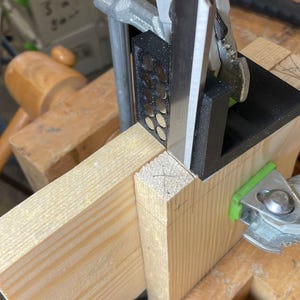 Peut inclure: Gros plan sur un projet de menuiserie avec deux morceaux de bois assemblés à l'aide d'une pince et d'une équerre en métal. Le bois est brun clair et la pince est verte et argentée.