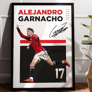 Könnte beinhalten: Gerahmtes Poster mit dem Fußballspieler Alejandro Garnacho in einem roten Trikot, mit seinem Namen und seiner Unterschrift. Das Poster hat einen schwarzen Rahmen und steht vor einer weißen Wand. Ein geflochtener Korb mit einer Pflanze steht daneben.
