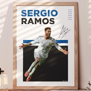 May include: Framed poster featuring Sergio Ramos in a dynamic pose, with the name in blue and black text. The image shows a soccer player in a white uniform against a black background. The poster is displayed on a wooden table.