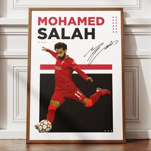 May include: Framed poster featuring Mohamed Salah in a red soccer uniform kicking a soccer ball. The poster has the name "MOHAMED SALAH" in large red and black letters. A signature is visible. The background is black and white.