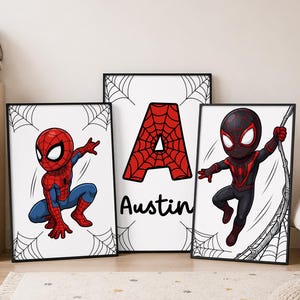 May include: Three framed wall art prints featuring a red and blue superhero in a crouching pose, a large red letter A with a web pattern above the name Austin, and a black and red superhero swinging on a web, all with spiderweb corner accents.