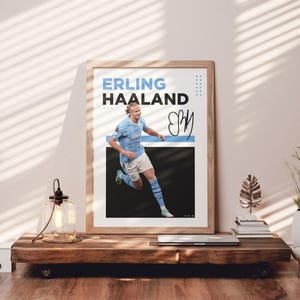 May include: Framed poster featuring Erling Haaland in a light blue soccer uniform, with the name "ERLING HAALAND" in blue and black text. The poster includes a signature and is displayed on a wooden table.