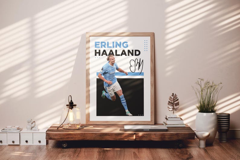 May include: Framed poster featuring Erling Haaland in a light blue soccer uniform, with the name "ERLING HAALAND" in blue and black text. The poster includes a signature and is displayed on a wooden table.