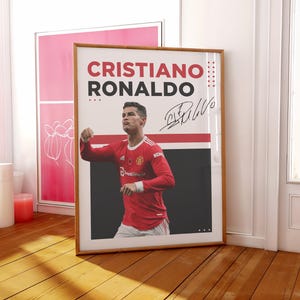 May include: A framed poster featuring Cristiano Ronaldo in a red soccer jersey. The poster has the name "CRISTIANO RONALDO" in large red and black letters, along with his signature. The image is set against a white and black background.
