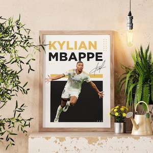 May include: Framed poster featuring Kylian Mbappe in a soccer uniform, with his name in bold yellow and black text. The poster includes a signature and is displayed on a shelf with plants and decorative items.