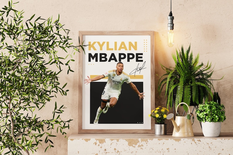 May include: Framed poster featuring Kylian Mbappe in a soccer uniform, with his name in bold yellow and black text. The poster includes a signature and is displayed on a shelf with plants and decorative items.