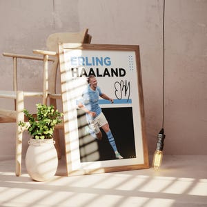May include: A framed poster featuring the name "ERLING HAALAND" in blue and white text, with a photo of a soccer player in a light blue uniform. The poster includes a signature and is displayed near a wooden chair and a plant.