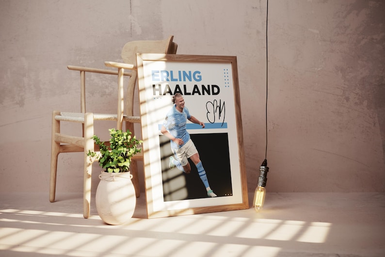 May include: A framed poster featuring the name "ERLING HAALAND" in blue and white text, with a photo of a soccer player in a light blue uniform. The poster includes a signature and is displayed near a wooden chair and a plant.