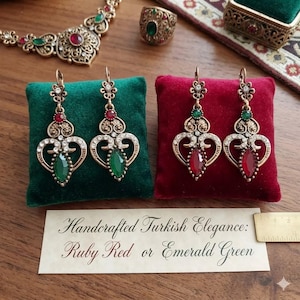 Könnte beinhalten: Verzierte goldfarbene Ohrringe mit herzförmigen Designs, besetzt mit klaren Kristallen und entweder smaragdgrünen oder rubinroten tropfenförmigen Edelsteinen. Die Ohrringe werden auf grünen und roten Samtkissen präsentiert. Eine passende Halskette und ein Ring sind ebenfalls sichtbar.