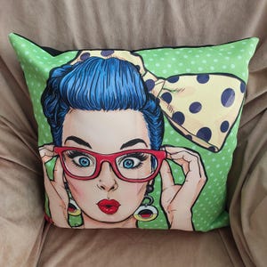 May include: Square decorative pillow featuring a pop art style illustration of a woman with blue hair, red glasses, and a yellow polka dot bow. The background is light green with white polka dots. The woman has red lips and is holding her glasses.