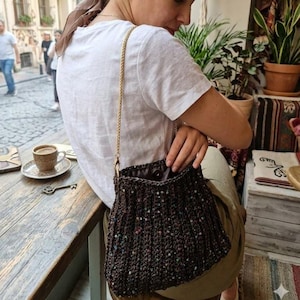 Può includere: Una borsa a mano all'uncinetto marrone scuro con una tracolla a catena dorata. La borsa è impreziosita da paillettes e ha un interno scuro. La borsa è tenuta da una persona che indossa una maglietta bianca.