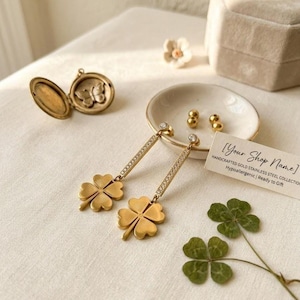 May include: Gold-colored clover earrings with a diamond-studded bar and a small, round diamond at the top. The earrings are displayed with a small dish of gold beads, a locket, and a four-leaf clover.