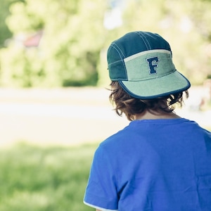 May include: A person wearing a blue t-shirt and a teal and green cap with the letter "F" on the front. The person is standing near a tree in a park-like setting with green grass and trees in the background.