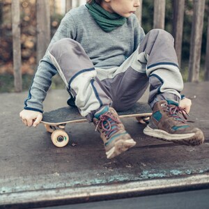 Pode incluir: Uma pessoa sentada numa prancha de skate. A prancha de skate é preta com rodas amarelas. A pessoa está vestindo calças cinzas com remendos cinza claro, uma camisa cinza de manga comprida e botas marrons e azuis. Um cachecol verde está em volta do pescoço.