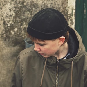 Puede incluir: Un gorro negro de pana. El sombrero se usa con una chaqueta con capucha verde oliva con cierre de cremallera y cordón. La chaqueta tiene un forro negro. El fondo es una pared texturizada de tonos neutros.