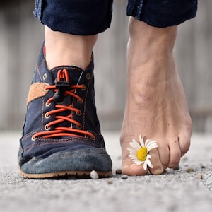 Op de afbeelding: Close-up van een marineblauwe en bruine schoen met oranje veters naast een blote voet met een madeliefje tussen de tenen. De schoen heeft een robuuste stijl en de veters zijn strak geregen. De foto is van een lage hoek genomen.