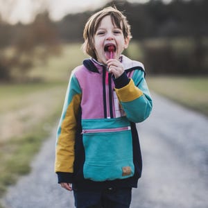 May include: A child wearing a colorful hooded sweatshirt with a teal front pocket, pink zipper, and yellow accents. The child is holding a red lollipop and standing on a gravel path.