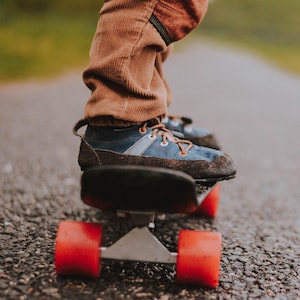 May include: A person's feet and skateboard are shown on a paved road. The skateboard has red wheels and a black deck. The person is wearing brown corduroy pants and blue and brown shoes with orange laces.